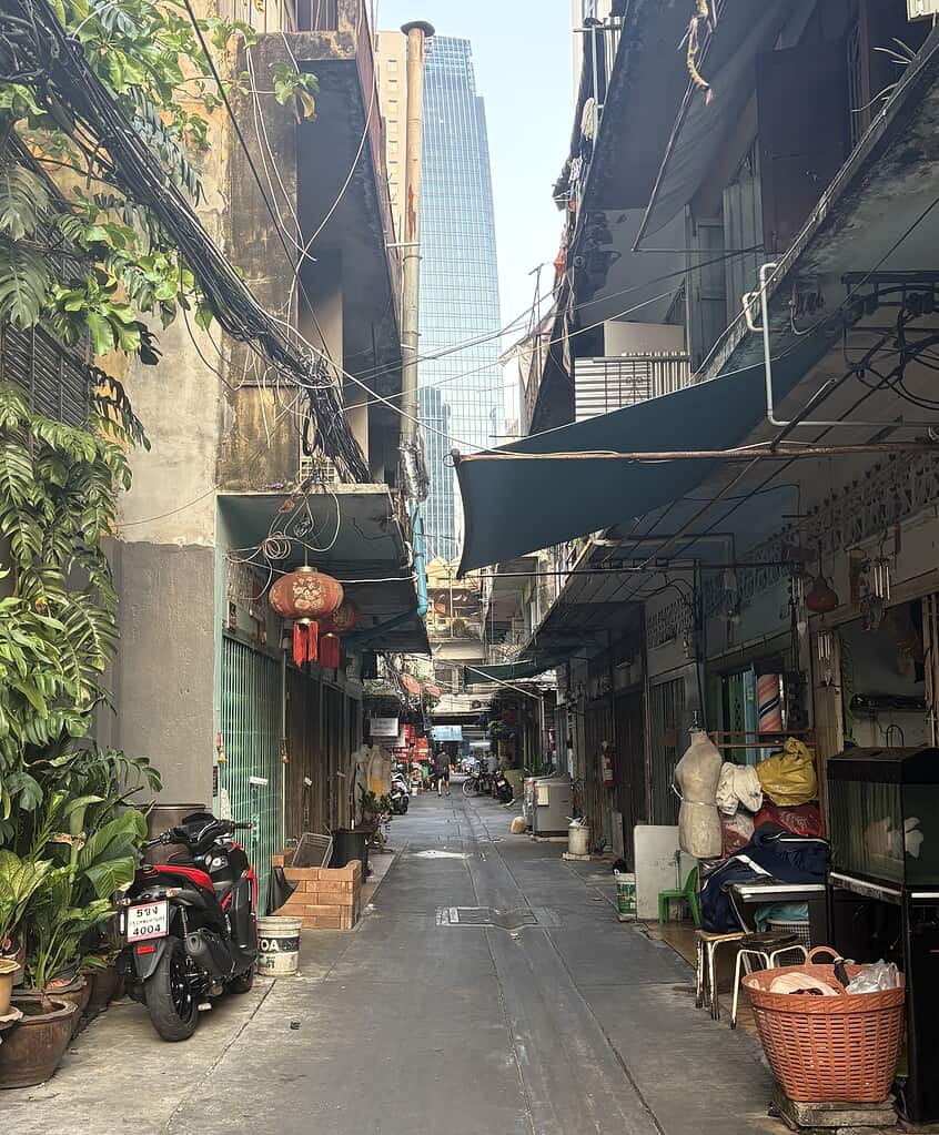 An alleyway in one of Bangkok’s lesser-visited neighborhoods, featuring old-school buildings and houses with a towering modern skyscraper in the background, capturing the city’s striking architectural contrast.