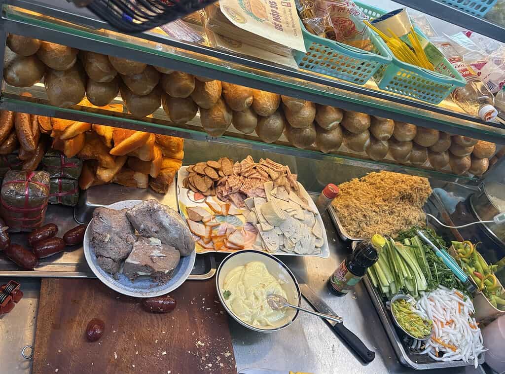 Close-up of a woman’s banh mi cart in Saigon showing stacked bread, cold cuts, and fresh vegetables on display.