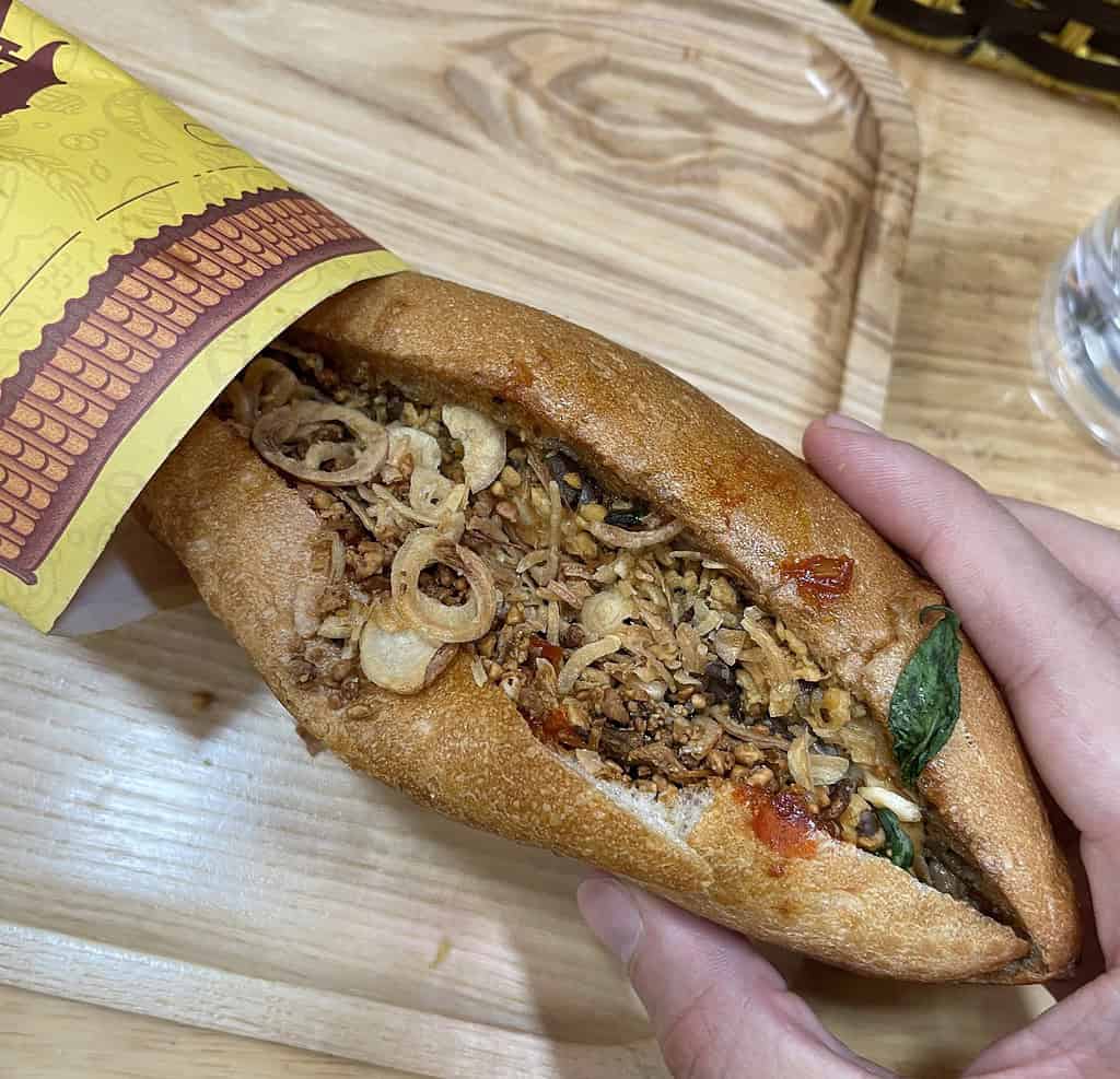 A close-up of a banh mi served in Hanoi, topped with crispy fried onions and fresh greens like mint.