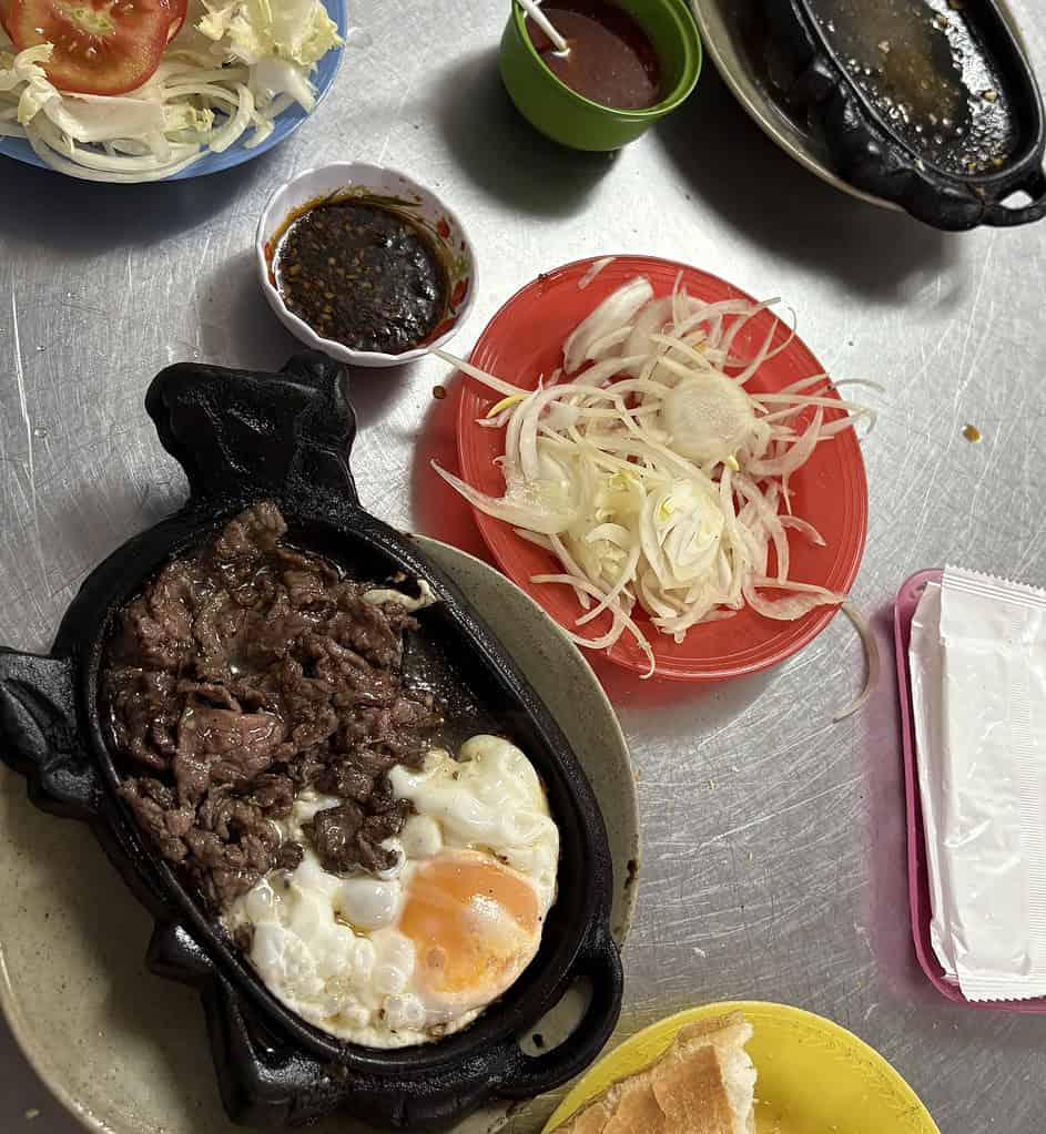 Close-up of Bo Ne sizzling on a hot plate in Saigon, served with raw onions, vegetables, and broken banh mi bread at one of the city’s top Bo Ne spots.