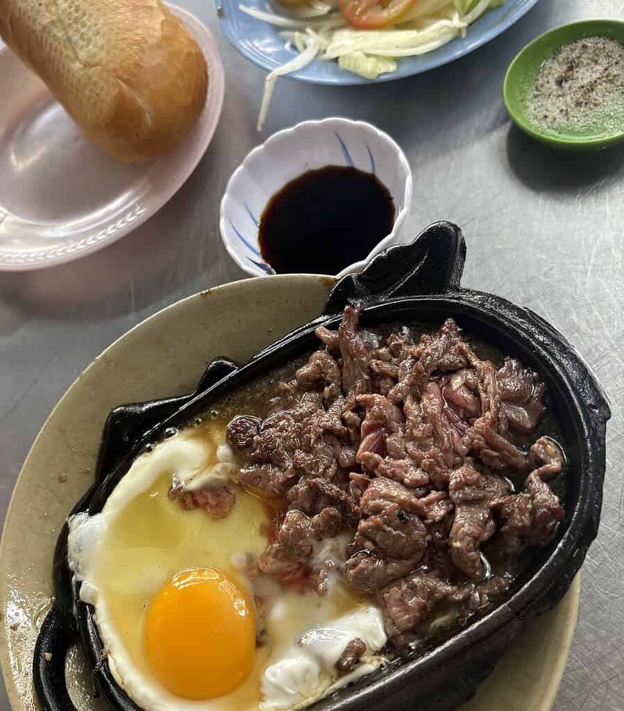 Close-up of sizzling medium-rare beef on a Bo Ne hot plate, served with banh mi bread, soy sauce, and a side of vegetables.
