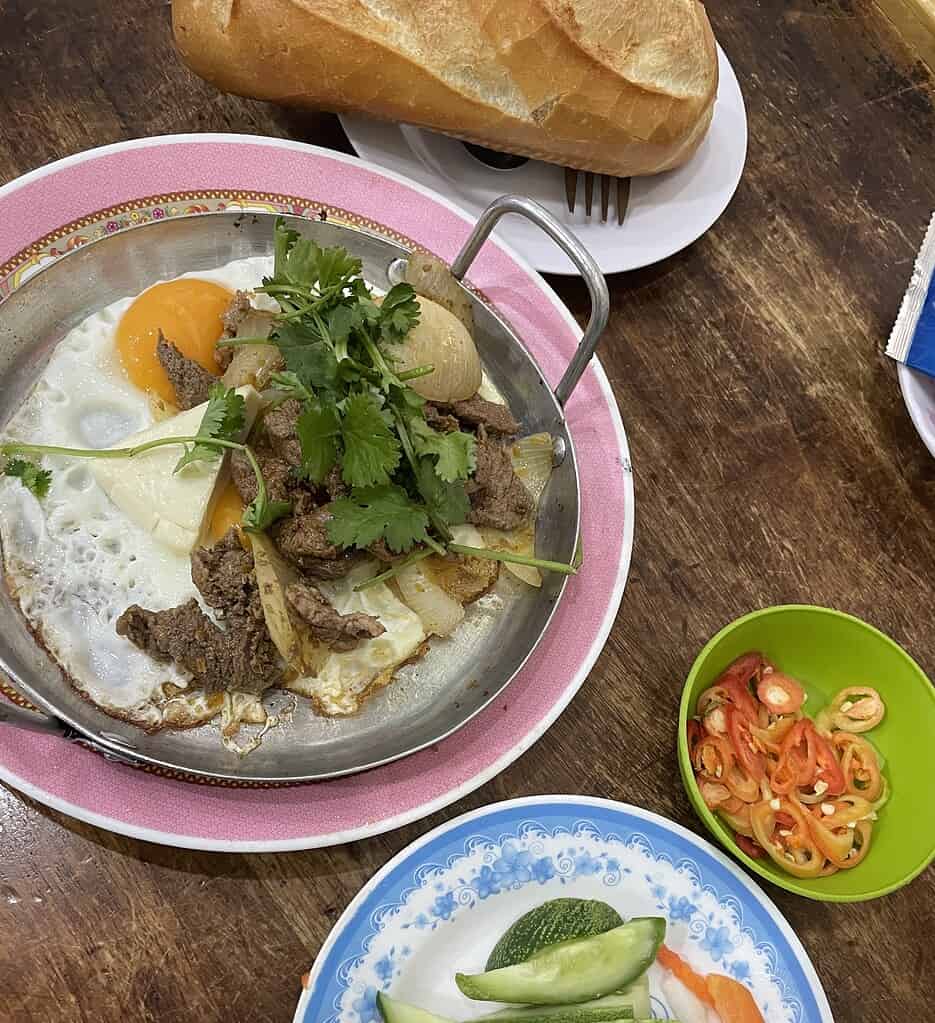 Bo Ne dish served with fried eggs, beef, fresh cilantro, a side of chilies, pickled vegetables, and crispy bread.