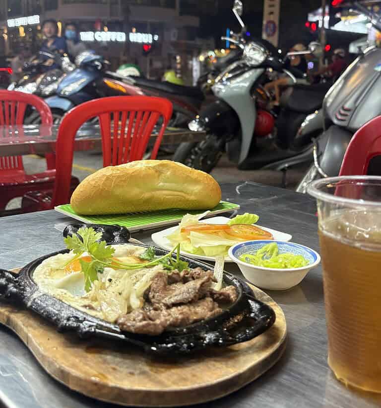 Street-side Bo Ne on a sizzling plate with Nuoc Sam and banh mi bread, with scooters and Saigon’s evening street scene in the background.