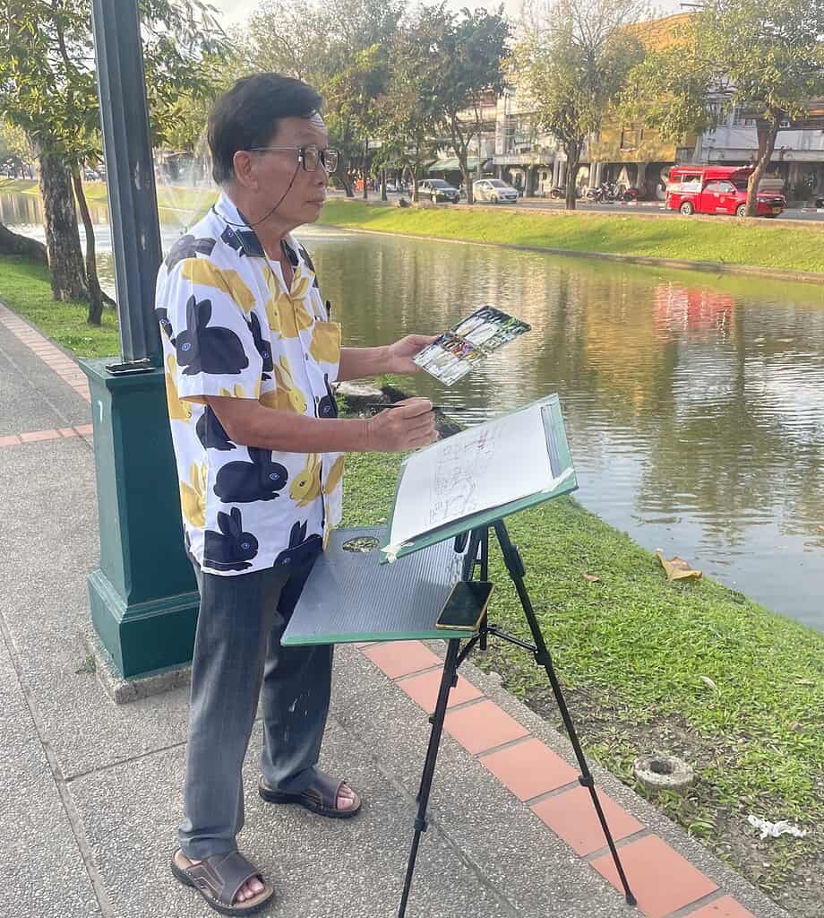 Local artist painting by the old city river in Chiang Mai, wearing glasses and working with his paint palette and easel.

