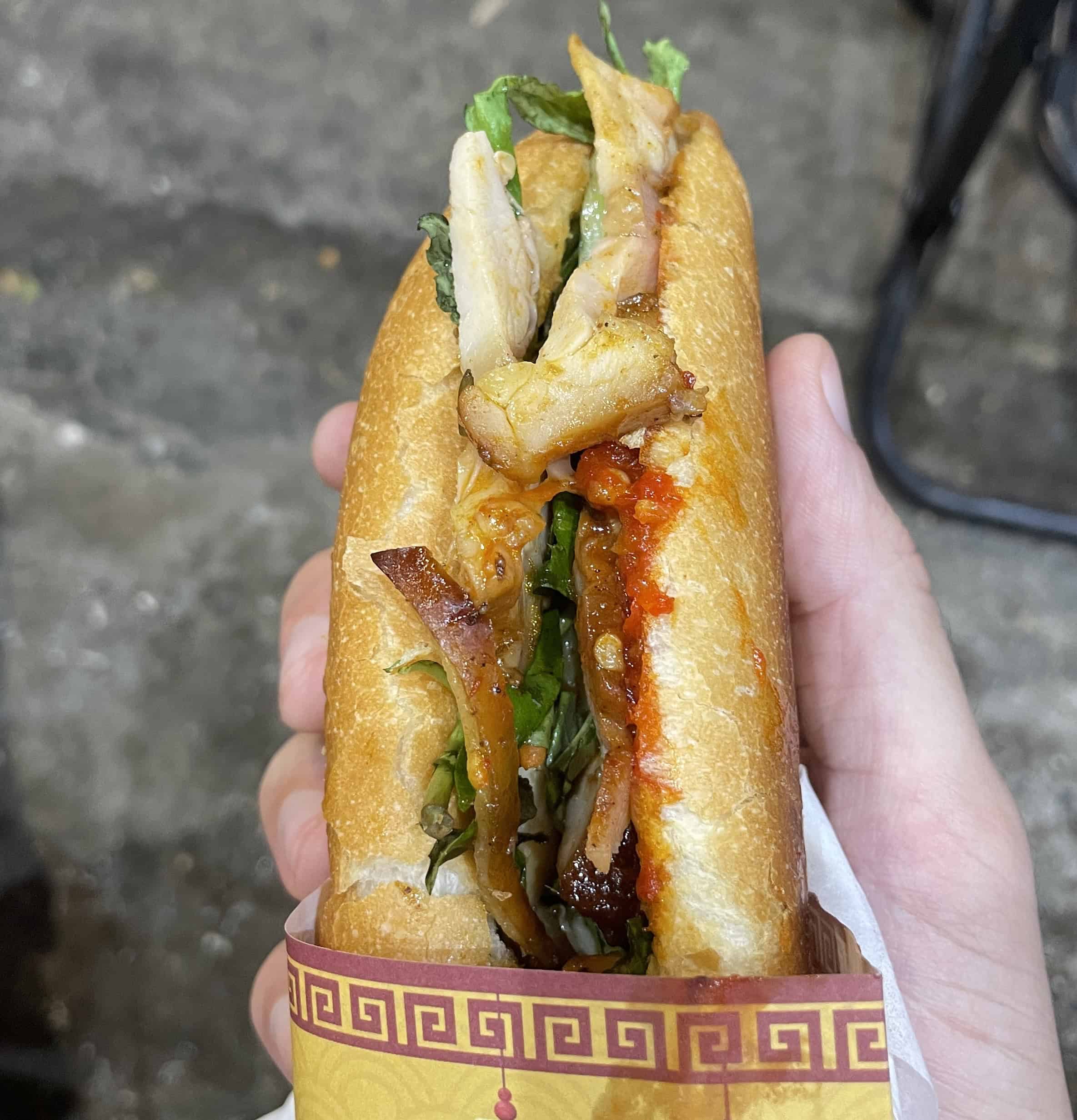 Closeup of a chicken banh mi in Hoi An, showing crispy bread, chili sauce, fresh vegetables, and sandwich wrapper underneath.