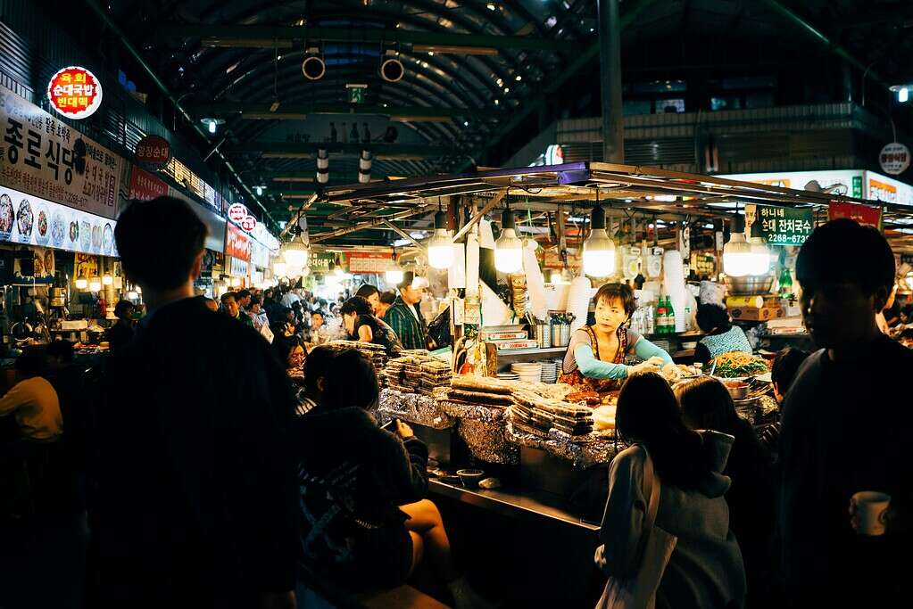 Nighttime scene at Gwangjang Market in Seoul with a vendor selling Korean street food to locals and tourists under bright lights.