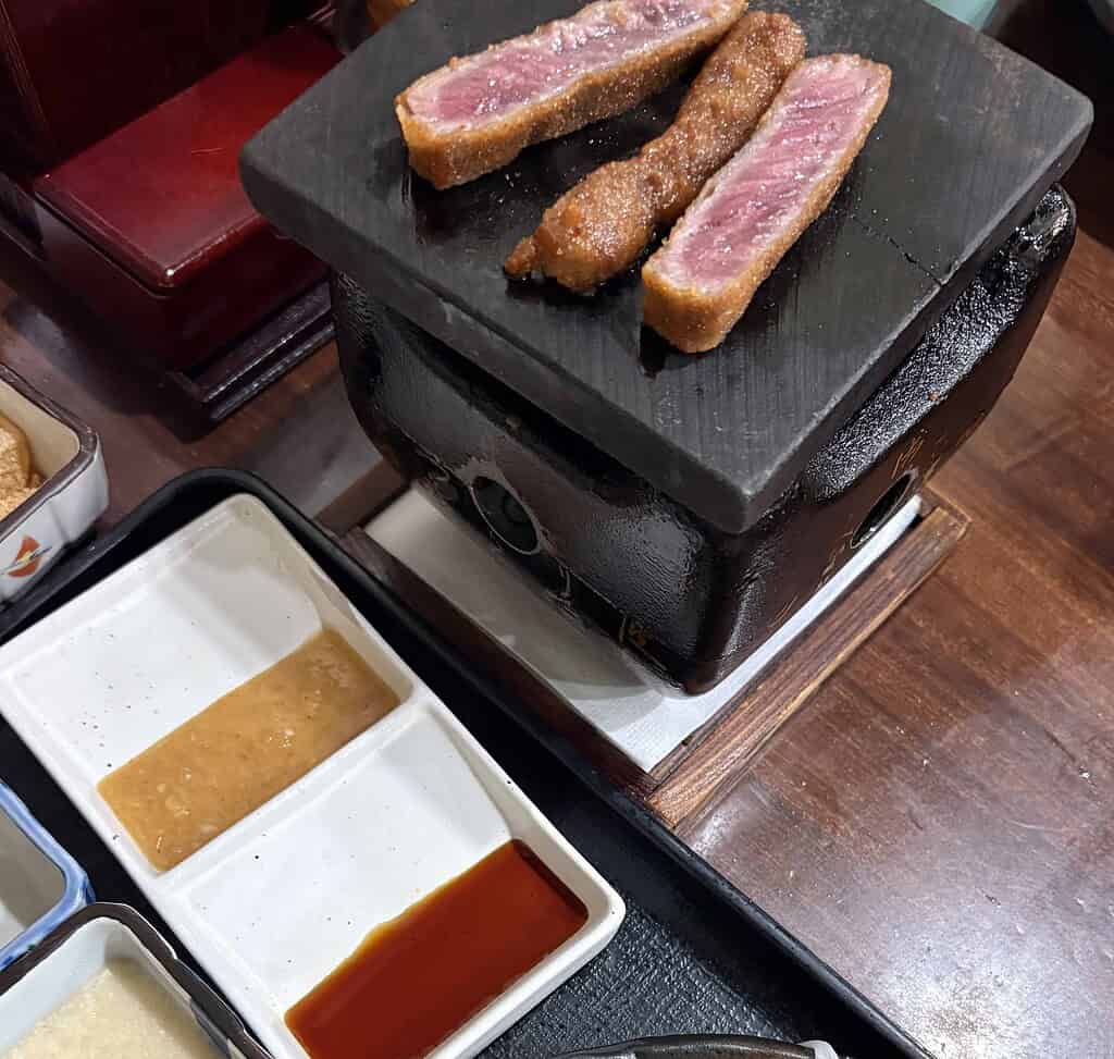 Beef cutlets sizzling on a hot stone grill at Gyukatsu Motomura, with flames underneath and dipping sauces nearby.