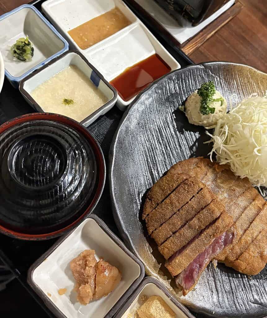 Close-up of a Gyukatsu Motomura set meal featuring miso soup, rice, dipping sauces, and an empty stone plate for grilling.