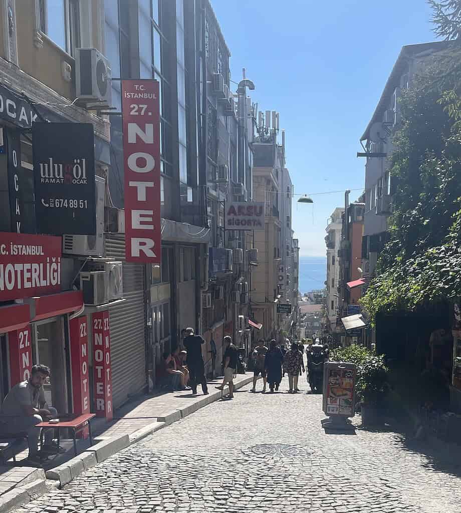 A hilly walkway in Istanbul lined with shops and cafés on both sides, with locals and tourists walking and a distant view of the river.