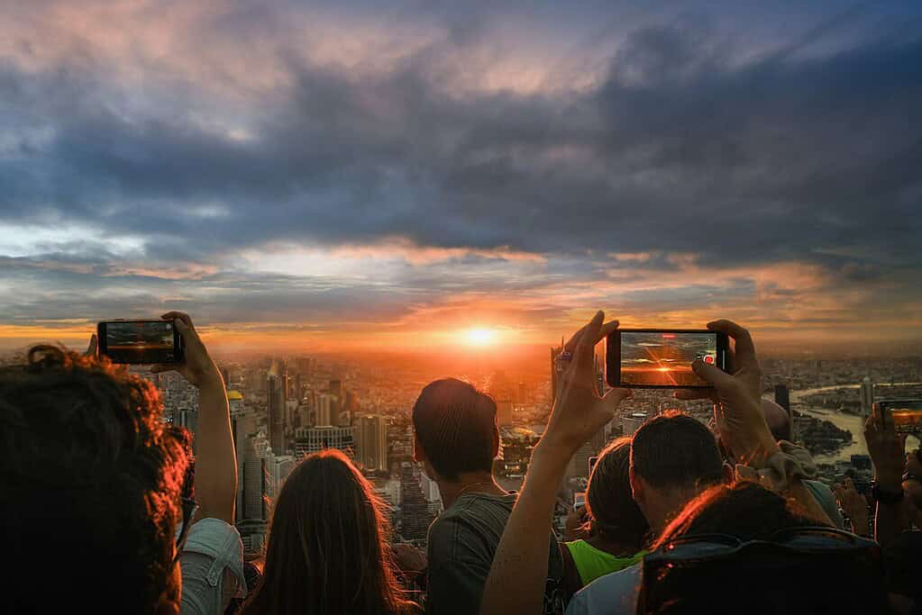 Crowd of people taking sunset photos from the top of the Mahanakhon Skywalk in Bangkok with panoramic city views.