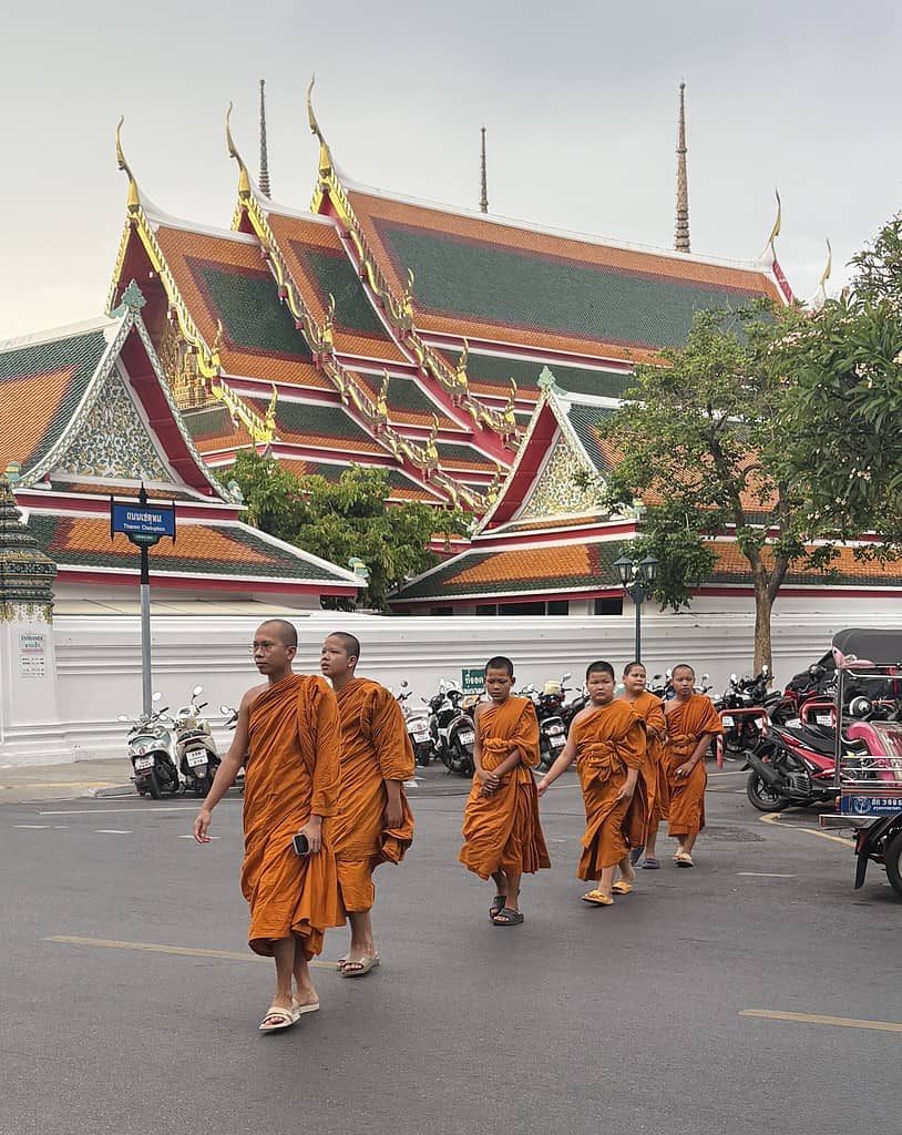 A senior monk leads a group of young monks in traditional saffron robes as they cross a quiet street in Bangkok, capturing a peaceful everyday moment in Thailand.