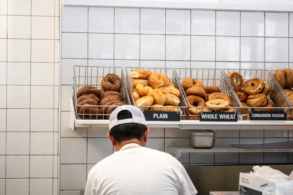 Here’s the revised alt text:📝 Alt Text:
A man preparing a bagel behind the counter of a New York City bagel shop, with a wide variety of bagels displayed in front of him.