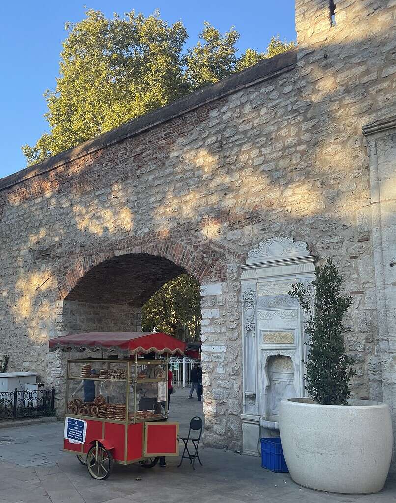 Old red simit cart in the Sirkeci area of Istanbul on a sunny afternoon, beside historic stone walls with a charming local vibe.
