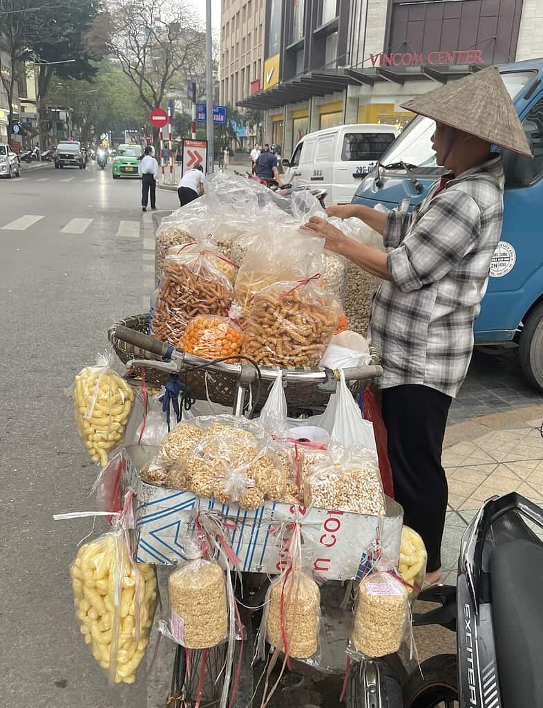 Street vendor in Hanoi, Vietnam selling a colorful variety of dried fruits and snacks from a sidewalk stall.
