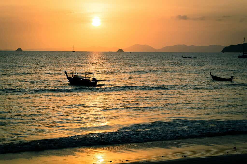 Sunset at Railay Beach in Krabi with longtail boats on the shore and towering limestone cliffs in the distance.