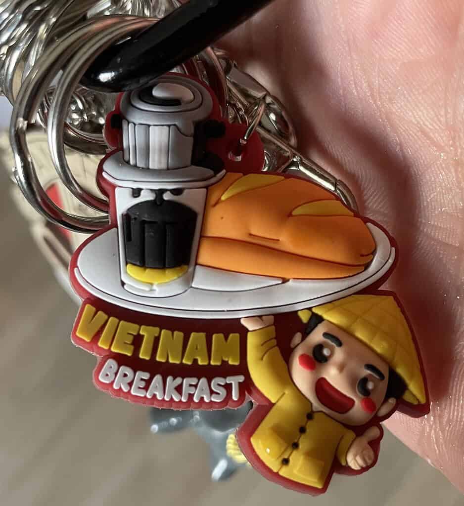 Cute Vietnamese keychain featuring a cartoon avatar and the phrase “Vietnamese Breakfast” with illustrations of banh mi and coffee.