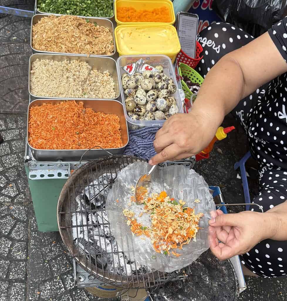 Woman in Saigon preparing Vietnamese street food known as “Vietnamese pizza” on rice paper with quail egg and toppings, grilled over charcoal.
