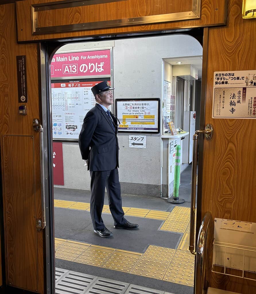Kyoto train worker in uniform standing on the platform, viewed from inside the train car with open doors during an Arashiyama day trip